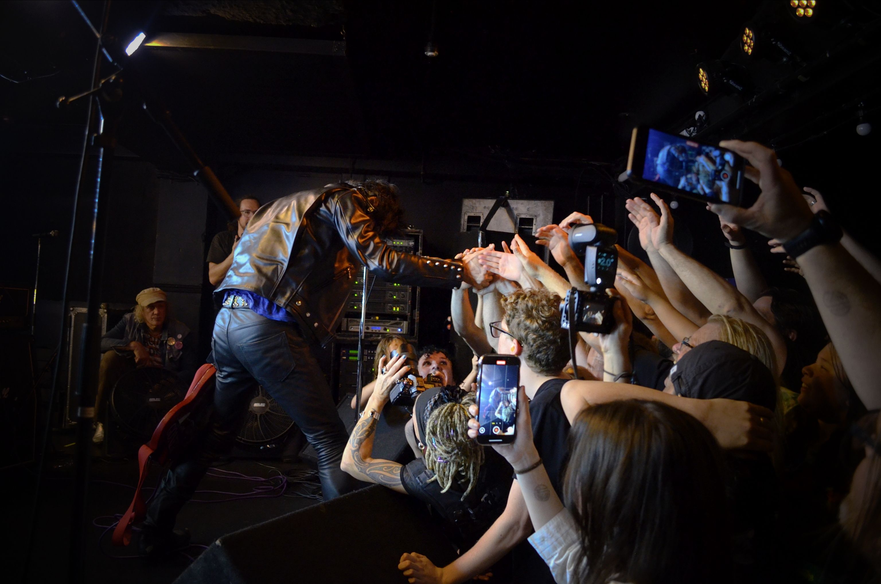 A chaotic image of a crowd reaching towards the frontperson of a band, who is facing away from us on the stage